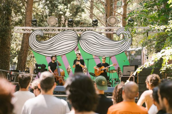 A band performs on an outdoor stage decorated with a black and white mustache design, with audience members watching in a wooded area.