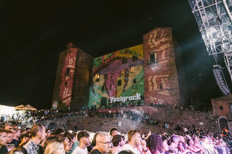 A large crowd of people attending an outdoor concert at night, with colorful projections on the ancient walls of a castle displaying the text 