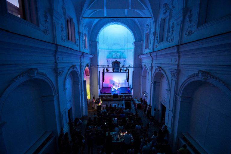 Interior of a church converted into a concert venue with atmospheric lighting and a crowd watching a performance on stage.