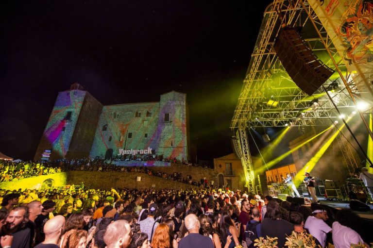 An outdoor music festival at night with a large crowd facing a lit stage, with light projections on a historic stone building as a backdrop.