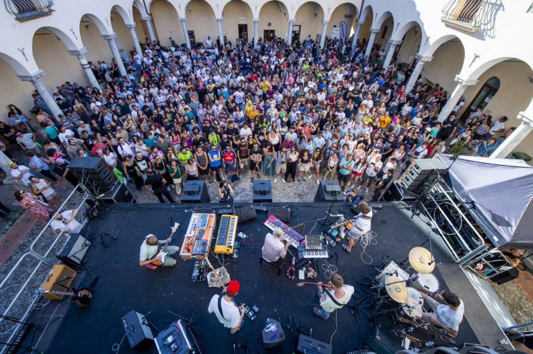 A band performs on an outdoor stage surrounded by a large crowd within an arched courtyard.