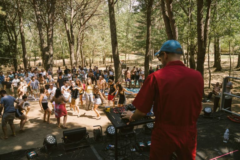 A DJ in a red outfit is operating equipment at an outdoor event, facing a crowd of people dancing and enjoying themselves in a wooded area.