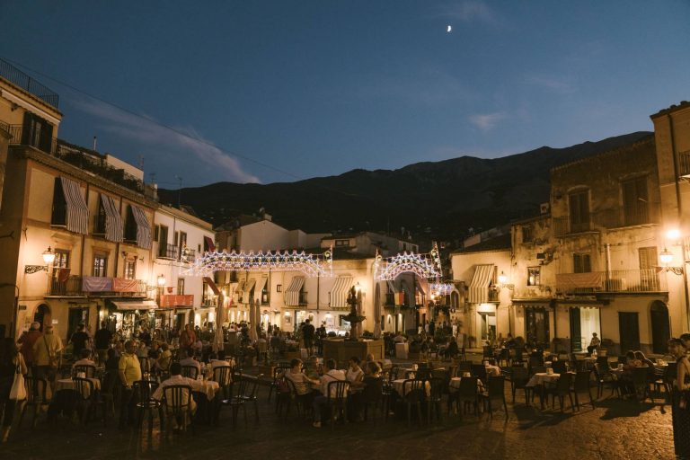 Outdoor evening scene at a bustling square filled with people dining at tables, with decorative lights strung above, traditional buildings, and a crescent moon in the sky.