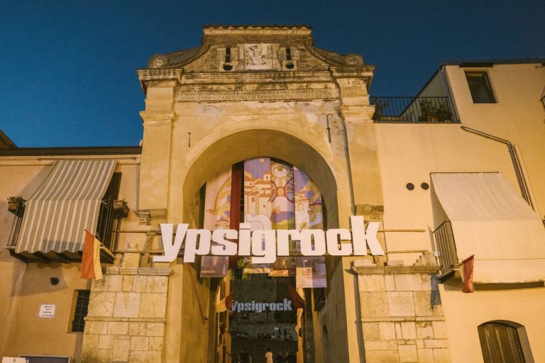 An old archway with the text "Ypsigrock" prominently displayed in the foreground against a twilight sky. A colorful mural is visible through the arch.