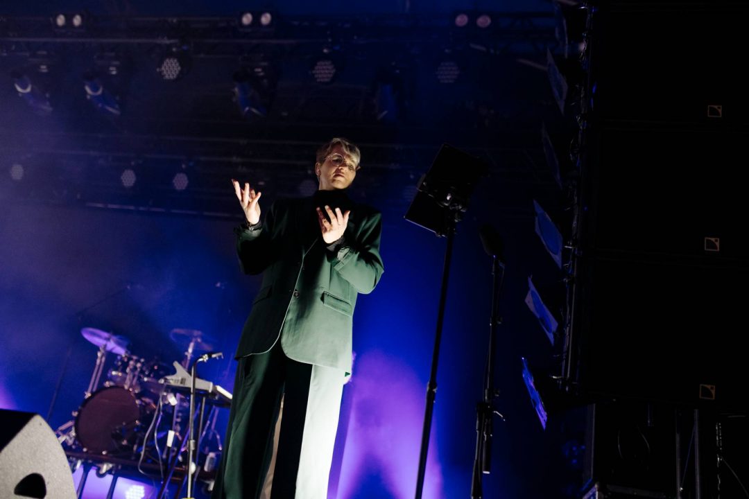 Man in green suit performing on stage with violet lighting in the background.