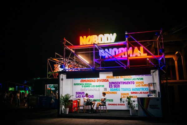 A nighttime scene of a vibrant outdoor event space with large glowing signs saying "NOBODY IS NORMAL" in colorful neon lights. The area is adorned with other messages about diversity and identity, and a few people are visible near the installation.