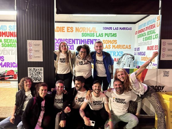 Group of nine people smiling and posing in front of a colorful booth with slogans advocating for sexual freedom and diversity, some wearing t-shirts with the text "NOBODY IS NORMAL".