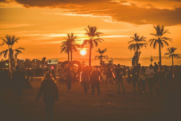 People walking at an outdoor event during sunset, with palm trees and bright orange skies in the background.