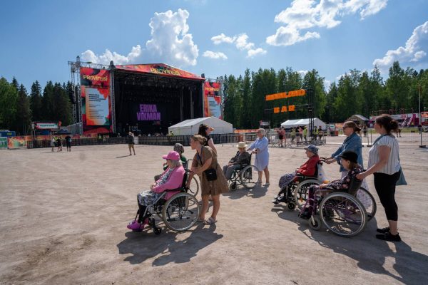 People in wheelchairs at an outdoor music festival with a stage in the background on a sunny day.