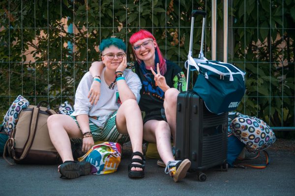 Two young people sitting on the ground leaning against a fence, one with teal hair and glasses, the other with pink hair, smiling and making a peace sign, surrounded by colorful luggage.