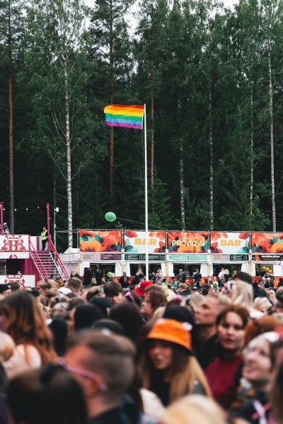 Crowded outdoor music festival with a colorful rainbow flag flying above the trees, people gathered near a bar area labeled "Provinssi."