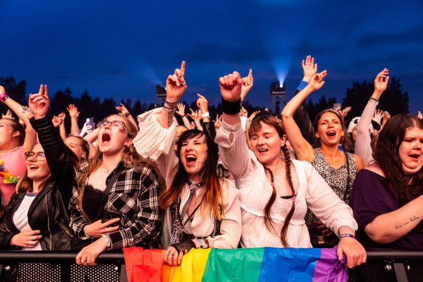 Excited crowd of people at a concert, cheering and singing along, with some holding a rainbow pride flag.