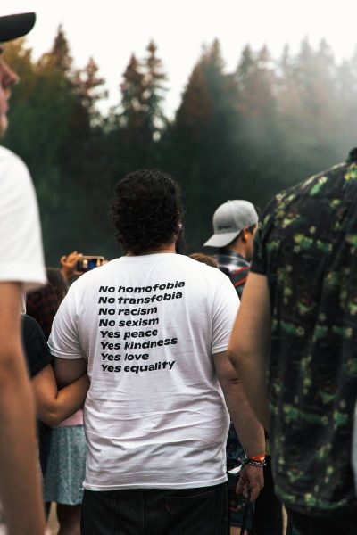 Person at an outdoor event wearing a white T-shirt with text advocating against homophobia, transphobia, racism, and sexism, and promoting peace, kindness, love, and equality. Other people and trees in the background.