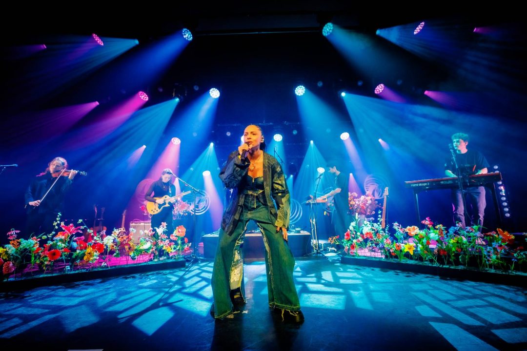 A female singer performing on stage with a band, surrounded by vibrant stage lighting and colorful flowers, with musicians playing violin, guitar, and keyboards.