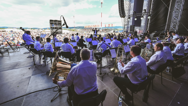 An orchestra performing on an outdoor stage with a large audience in the background.