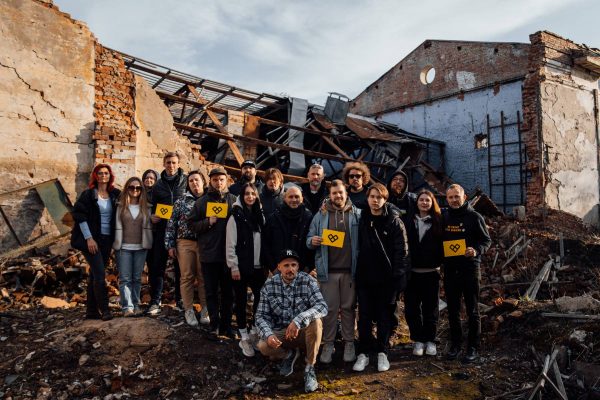 The Music Ambassadors Tour 2024 group standing in front of a destroyed building in Ukraine, with some individuals holding the yellow Take a Stand signs featuring a black heart symbol.