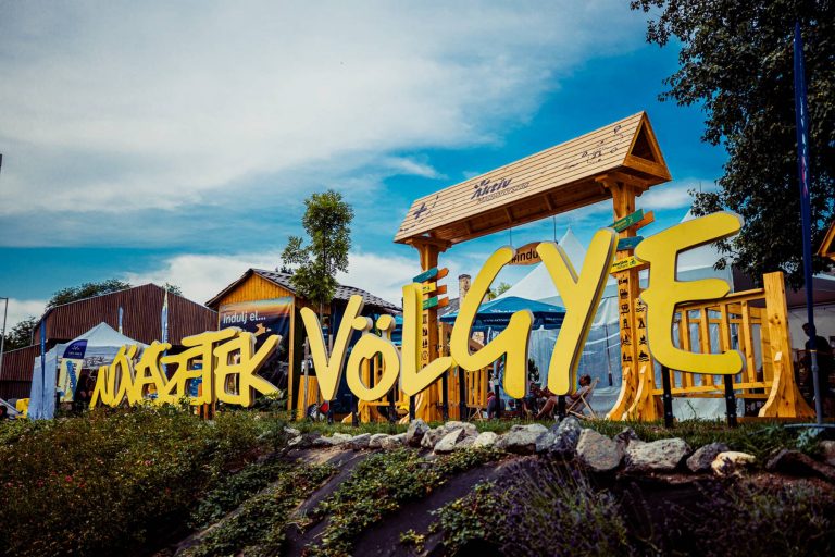 Large yellow "Művészetek Völgye" sign with a wooden structure and tents in the background on a sunny day.