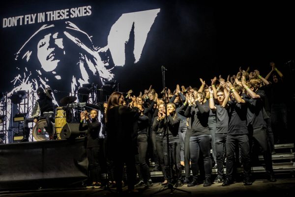 A large choir dressed in black performs on stage with a black and white image and text "DON'T FLY IN THESE SKIES" displayed on the screen behind them.