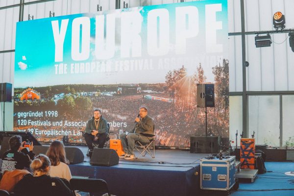 Two people sitting on a stage in front of a large screen displaying information about a European festival association.