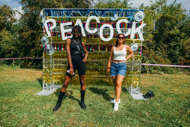 Two people posing in front of a colorful "PEACOCK" backdrop at an outdoor event.