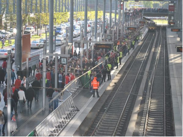 Crowded train station platform with people waiting near the tracks, security personnel in high-visibility vests present, and multiple signs visible.
