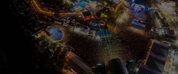 Aerial view of a large, brightly lit music festival at night, featuring stages with colorful lighting, a ferris wheel, tents, and a massive crowd of attendees.