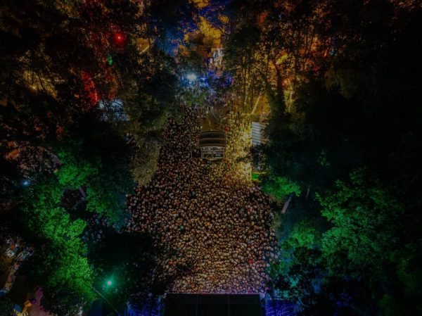 Aerial view of a large crowd gathered in a park-like setting during nighttime, surrounded by illuminated trees and colorful lights, with a stage and equipment visible.