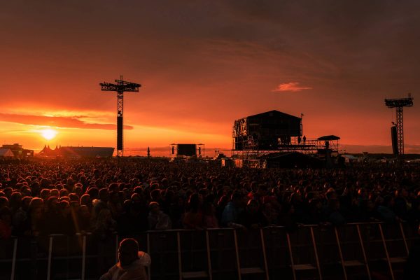 A large crowd gathered at an outdoor concert during sunset, with the sky lit in warm orange hues and several large stage structures silhouetted against the horizon.