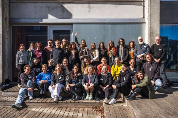 Large group of diverse people posing together outside on wooden steps in front of a modern building, some sitting and some standing, with a sunny and clear sky.