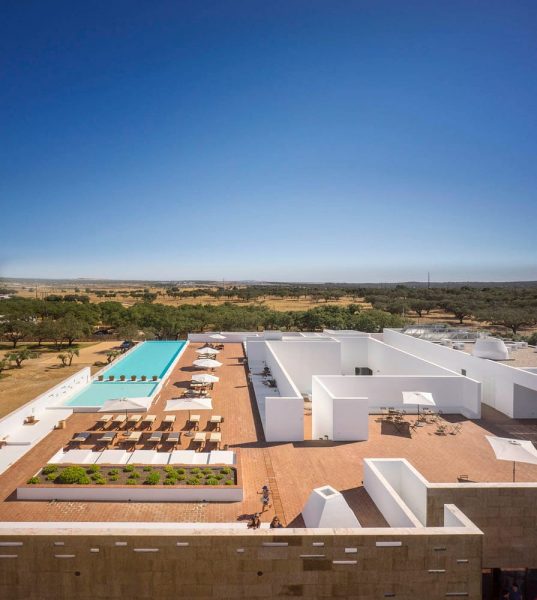A rooftop view of a modern building with white walls and brick flooring, featuring a rectangular swimming pool surrounded by lounge chairs and umbrellas, set against a backdrop of sparse trees and a clear blue sky.