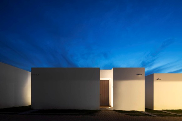 Modern minimalist building facade with flat, white walls and a single illuminated door at dusk under a deep blue sky