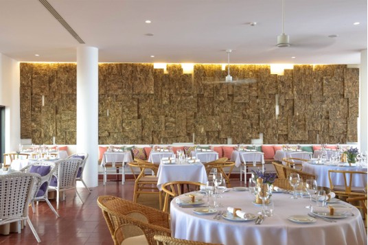 Bright and airy restaurant interior with white tablecloth-covered tables set with plates, glasses, and cutlery, featuring a mix of wicker and woven chairs, a textured wood-paneled wall with warm backlighting, and a cushioned bench along the wall.