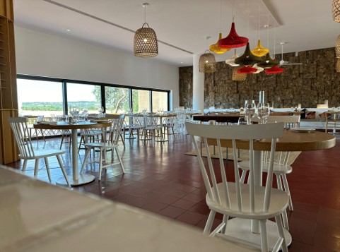 Spacious restaurant interior with white wooden chairs and round tables, red and yellow hanging pendant lights, large windows allowing natural light, and a textured stone wall in the background