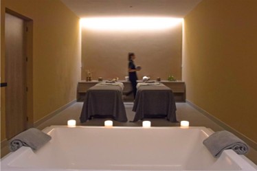 Spa room with two massage tables covered in gray sheets, a row of lit candles in front, gray towels placed on a white bathtub edge, and a person walking in the background against a softly lit wall