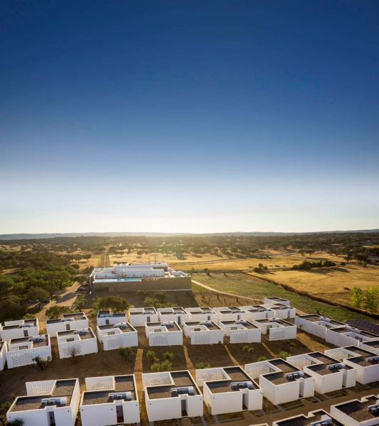 Aerial view of a modern housing complex with white, box-shaped buildings cast in long shadows under a clear blue sky, surrounded by dry grassy fields and scattered trees.