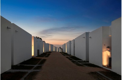 A pathway lined with modern white buildings on both sides during sunset or dusk, with warm light illuminating some entrances and a clear blue sky with some clouds above.