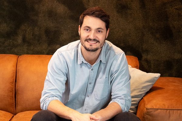 Man with dark hair and beard wearing a light blue button-up shirt sitting on a brown leather couch with a beige pillow against a dark textured background