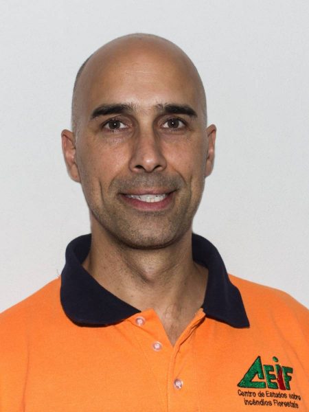 Portrait of a bald man wearing an orange polo shirt with a black collar and a logo on the left chest reading 'Centro de Estudios sobre Incendios Forestales'.