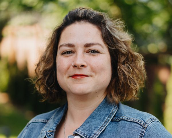 Close-up portrait of a woman with curly brown hair wearing a denim jacket and a nose ring, standing outdoors with a blurred green background