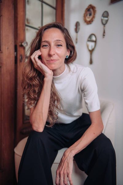 A woman with long wavy hair wearing a white short-sleeve top and black pants sits on a white chair, resting her head on her hand and smiling slightly, with decorative hand mirrors and a wooden door in the background.