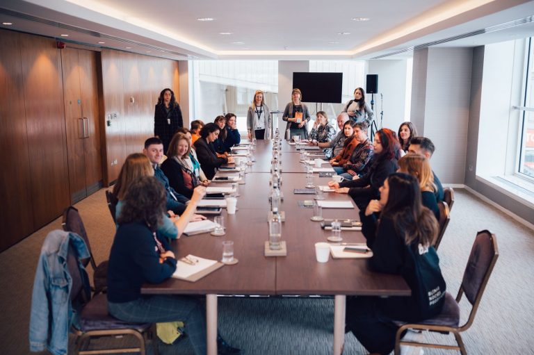 A large group of people seated around a long conference table in a modern meeting room, with three individuals standing at the far end near a screen and speakers, engaged in a discussion or presentation.