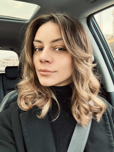 Woman with light brown wavy hair wearing a black turtleneck and jacket, sitting in a car with a seatbelt fastened, looking directly at the camera with a slight smile.
