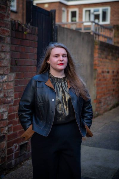 Young woman with long light brown hair wearing a black leather jacket and shiny dark blouse standing outdoors against a brick wall background.