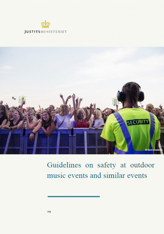 A crowd of people at an outdoor music event, cheering near the front barrier, with a security guard wearing a bright yellow vest and headphones facing them under a clear sky.