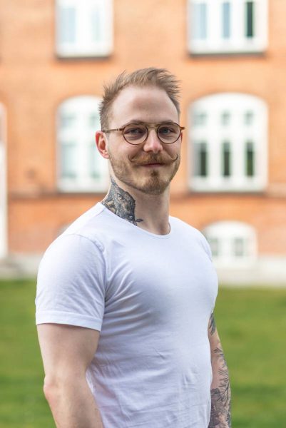 A man with glasses, a curled mustache, neck and forearm tattoos wearing a white t-shirt, standing outdoors with a blurred brick building in the background.