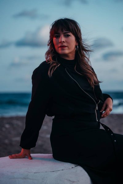 Young woman with shoulder-length wavy hair wearing a black outfit and silver hoop earrings sitting on a white surface at the beach during dusk with a cloudy sky and ocean in the background.