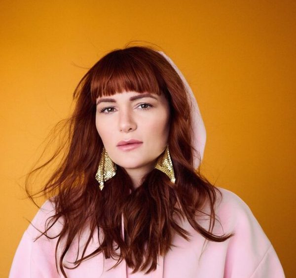 Woman with long red hair and bangs wearing a pink hoodie and large gold earrings against an orange background