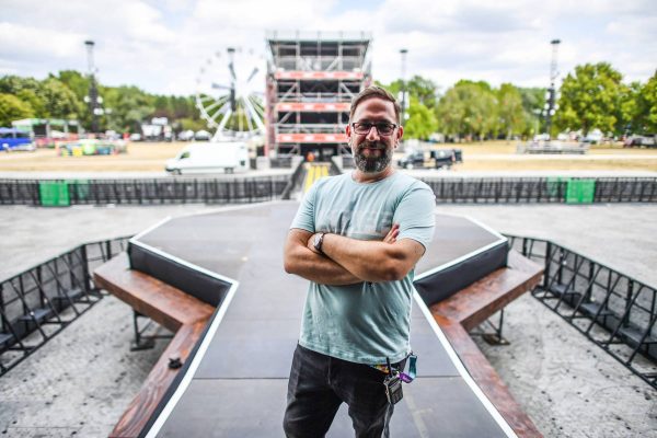 Man with glasses and beard standing confidently with arms crossed on an empty outdoor concert stage, with event scaffolding and a ferris wheel visible in the background under a cloudy sky.