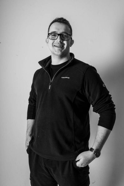 Black and white photo of a smiling man wearing glasses, a zip-up sweater, and a wristwatch, with hands resting in his pockets, standing against a plain wall.
