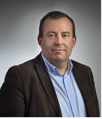 Portrait of a middle-aged man with short dark hair wearing a blue shirt and a dark brown jacket, against a neutral gray background.